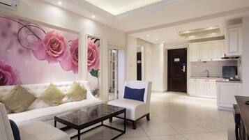 A modern living room with a white sofa, a coffee table, and a chair with a blue cushion. There is a large floral mural on the wall. The room leads to a kitchen with white cabinets and a tiled backsplash.