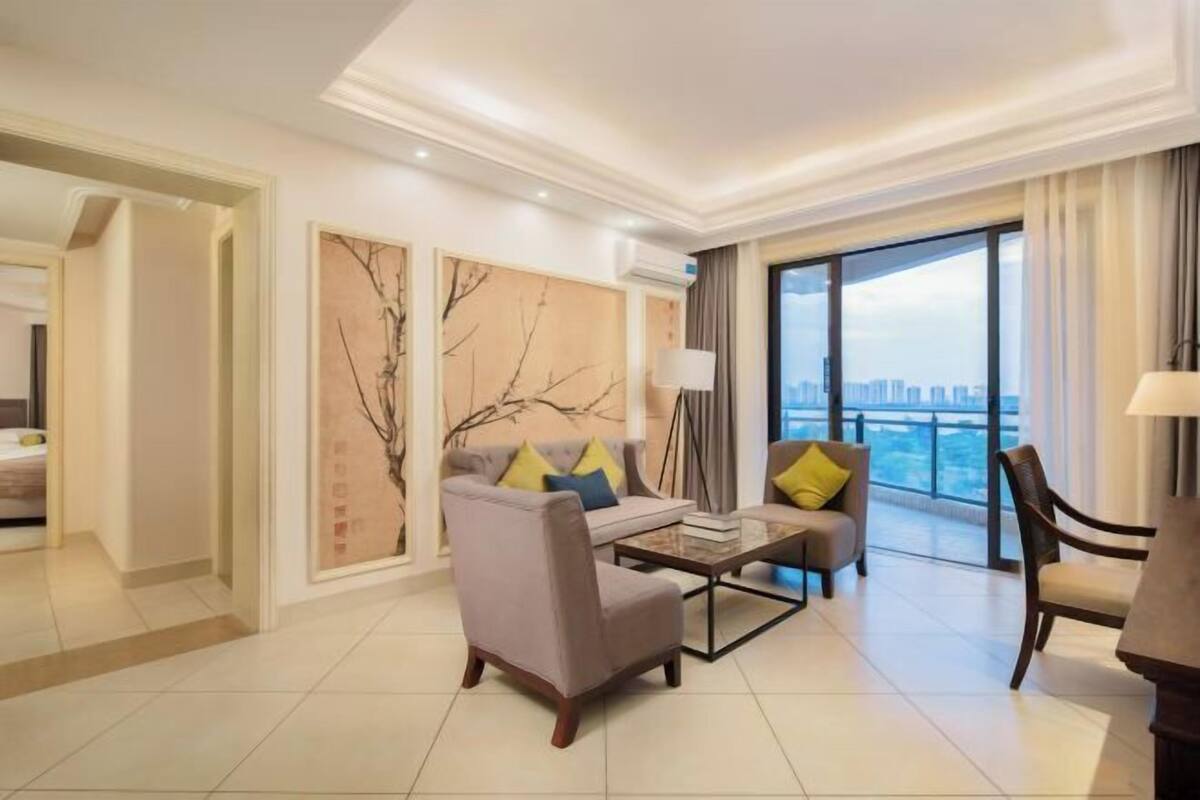 A modern hotel room with a sofa, armchair, and a coffee table. There is a large window offering a city view.