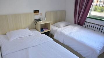 A hotel room with two single beds, a bedside table with a phone and a small shelf, and a window with curtains.