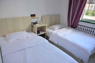 Habitación de hotel con dos camas individuales, una mesita de noche con un teléfono y un estante pequeño, y una ventana con cortinas.