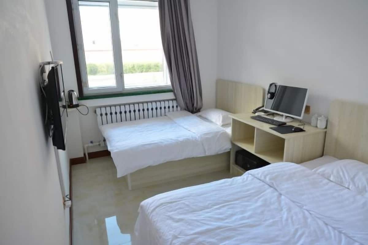 Habitación de hotel con dos camas individuales, un escritorio con computadora, un radiador y una ventana con cortinas.