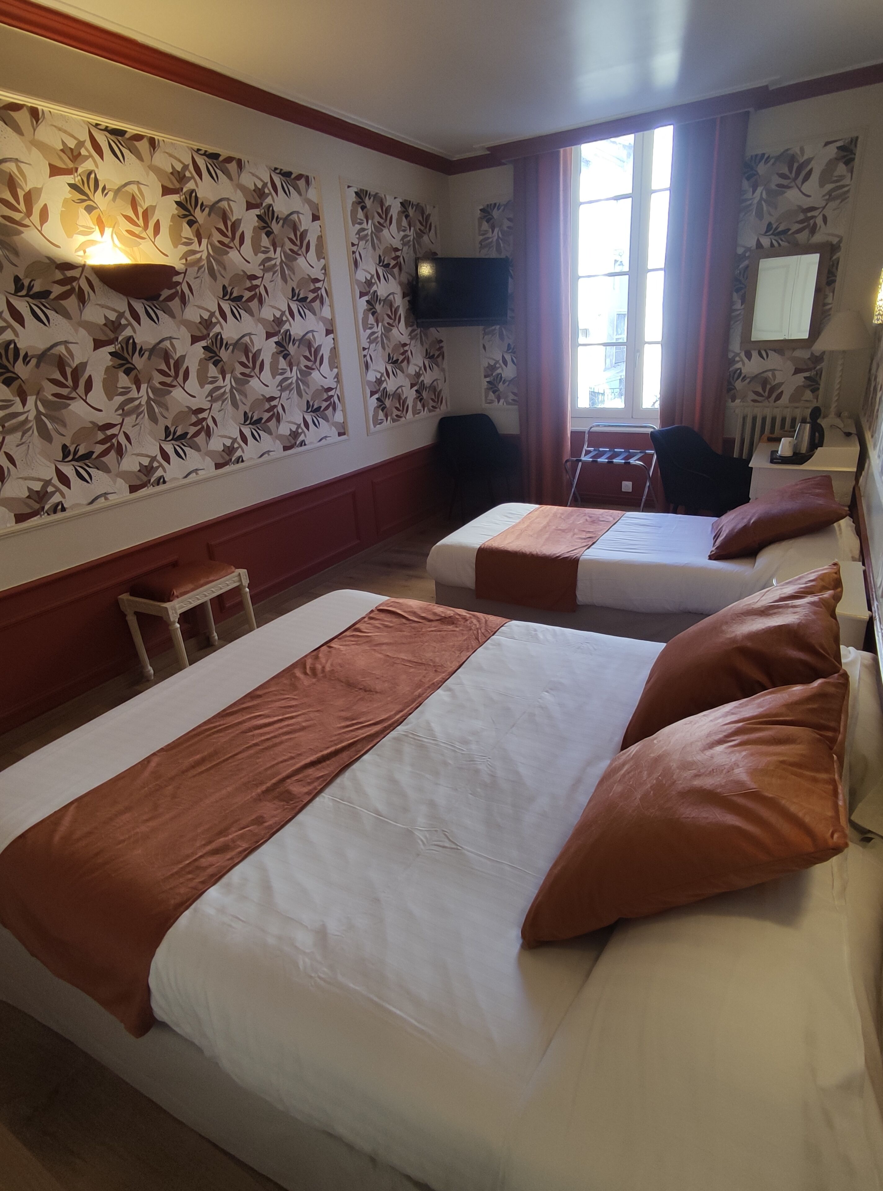 A hotel room with two beds, a television mounted on the wall, a window with curtains, and a radiator.