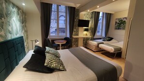 Quarto de hotel moderno com uma cama grande, uma escrivaninha com televisão e vista para edifícios históricos de pedra pelas janelas.