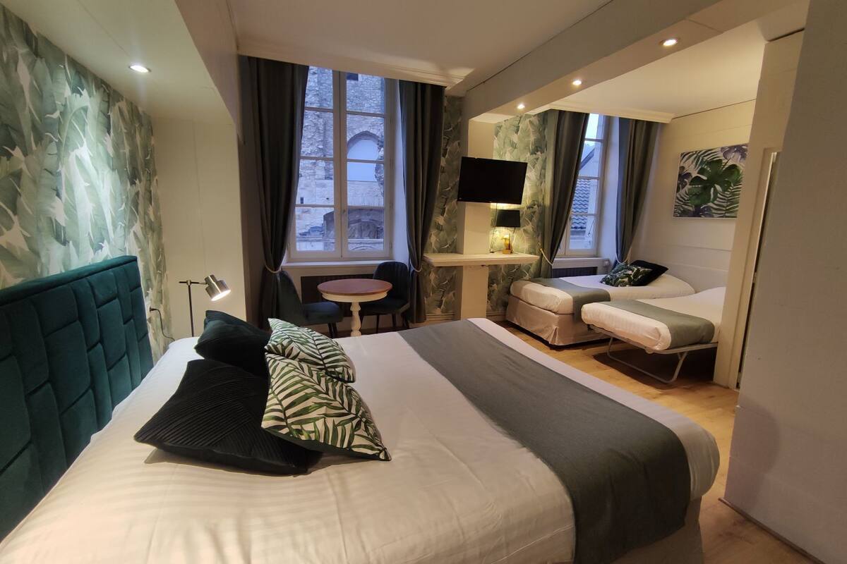 A modern hotel room with a large bed, a desk with a TV, and a view of historic stone buildings through the windows.