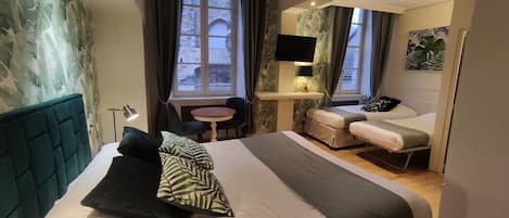 Quarto de hotel moderno com uma cama grande, uma escrivaninha com televisão e vista para edifícios históricos de pedra pelas janelas.