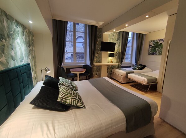 A modern hotel room with a large bed, a desk with a TV, and a view of historic stone buildings through the windows. - Le Pavillon Saint Martin (Souillac)