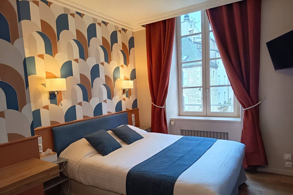 A hotel room with a bed, a nightstand, a wall-mounted TV, and a window with curtains.