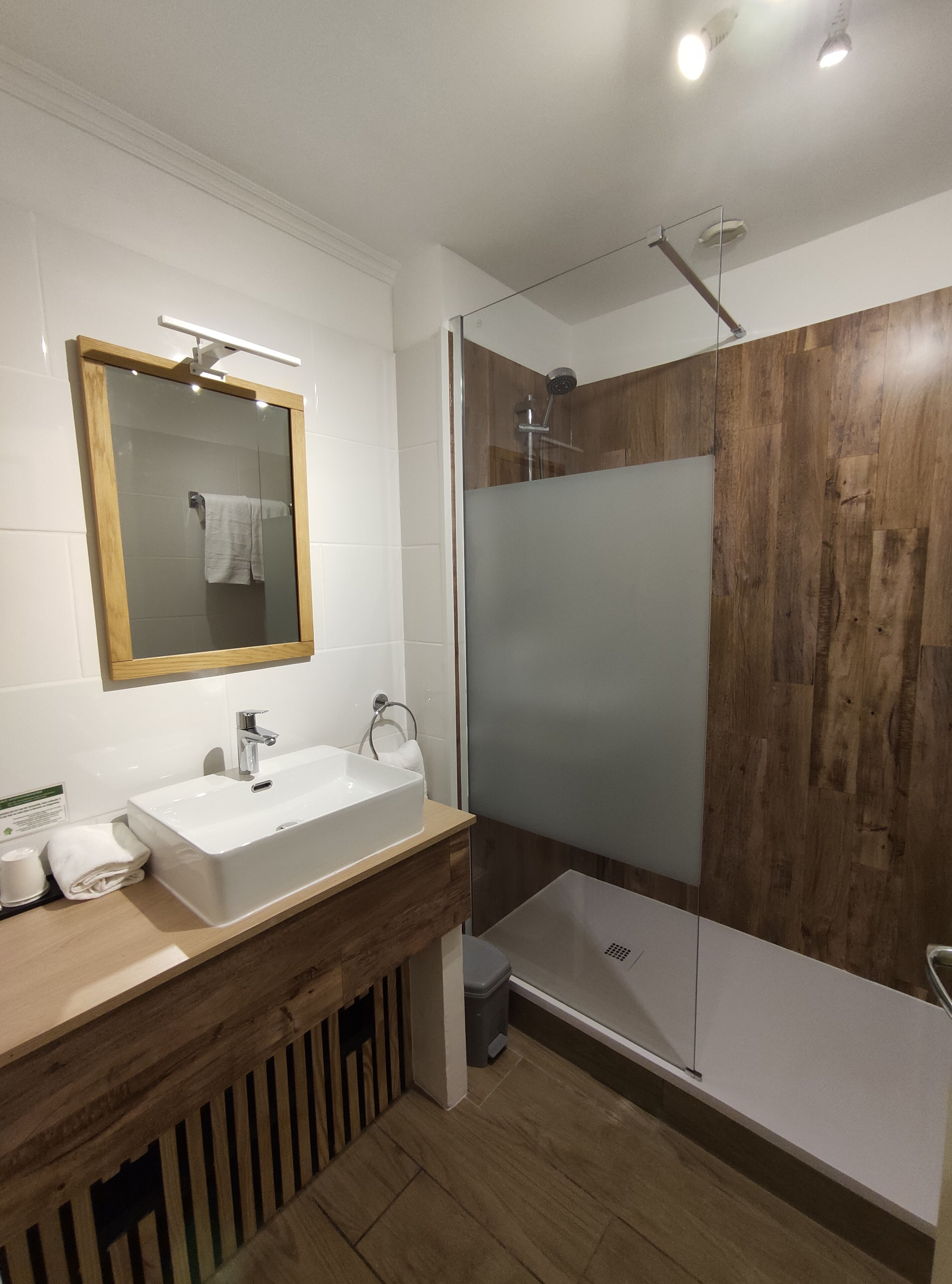 A modern bathroom with a wooden vanity, a large mirror, a white sink, and a glass-enclosed shower.