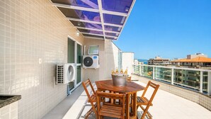 A rooftop terrace with a wooden table and chairs, air conditioning units, and a glass balustrade overlooking a cityscape. - Cf02 Cobertura 200m da Praia das Dunas (Cabo Frio)