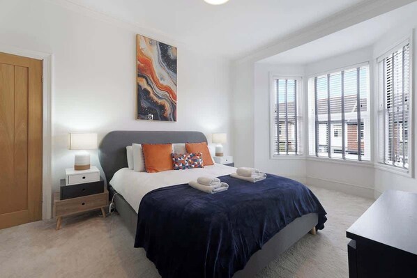 Une chambre à coucher comprenant un lit, des tables de chevet, une commode, une fenêtre avec des stores et un tableau accroché au mur.