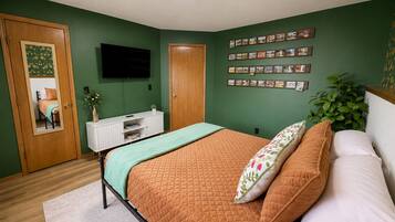 A bedroom with a bed, a TV, a white console, a plant, and a wall with pictures.
