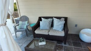 A covered patio area with a wicker sofa, a chair, a small table with a plant, and a white outdoor toilet.
