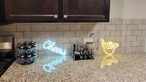 A kitchen with dark wooden cabinets, a granite countertop, and neon signs spelling out "Cheers" and "Good."