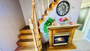 Una escalera con barandillas de madera, un reloj en la pared, una chimenea encendida y una mesa pequeña con adornos.