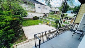 Un balcón residencial con barandilla de metal, un pequeño espacio de descanso y vistas a casas vecinas y vegetación.