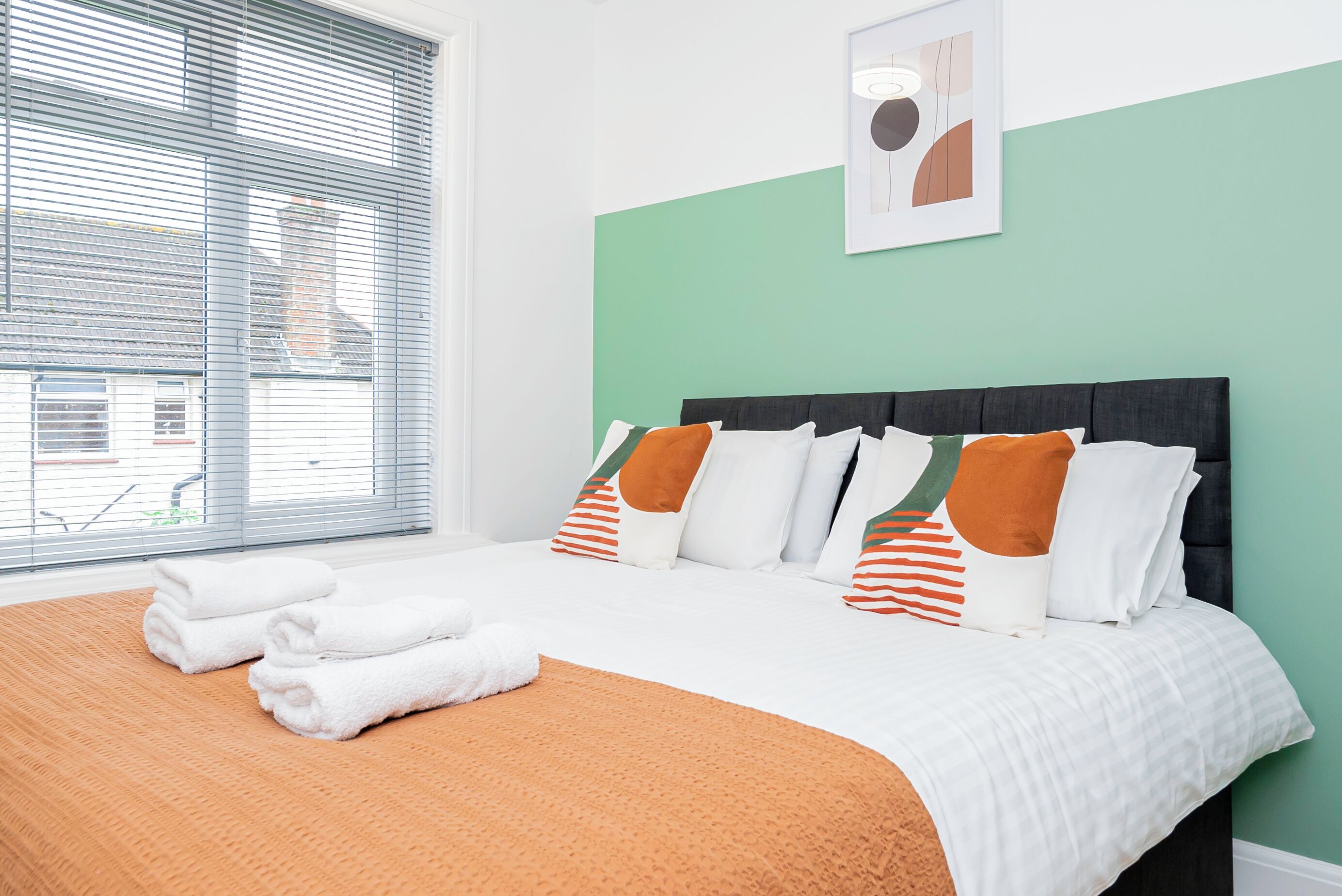 A neatly made bed with white and orange bedding, a green wall, and a framed picture above it.