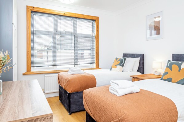 Een hotelkamer met twee bedden, een houten tafel, een raam met zonwering en een lijst met een afbeelding aan de muur.