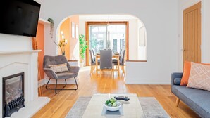 A modern living room with a fireplace, a flat-screen TV, a dining area with a table and chairs, and a sofa with cushions.