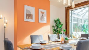 A dining area with a wooden table set for four, framed artwork on the wall, and a large window with a view of greenery.