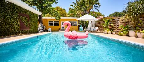 A swimming pool with a pink flamingo float, surrounded by lush greenery and a yellow building.