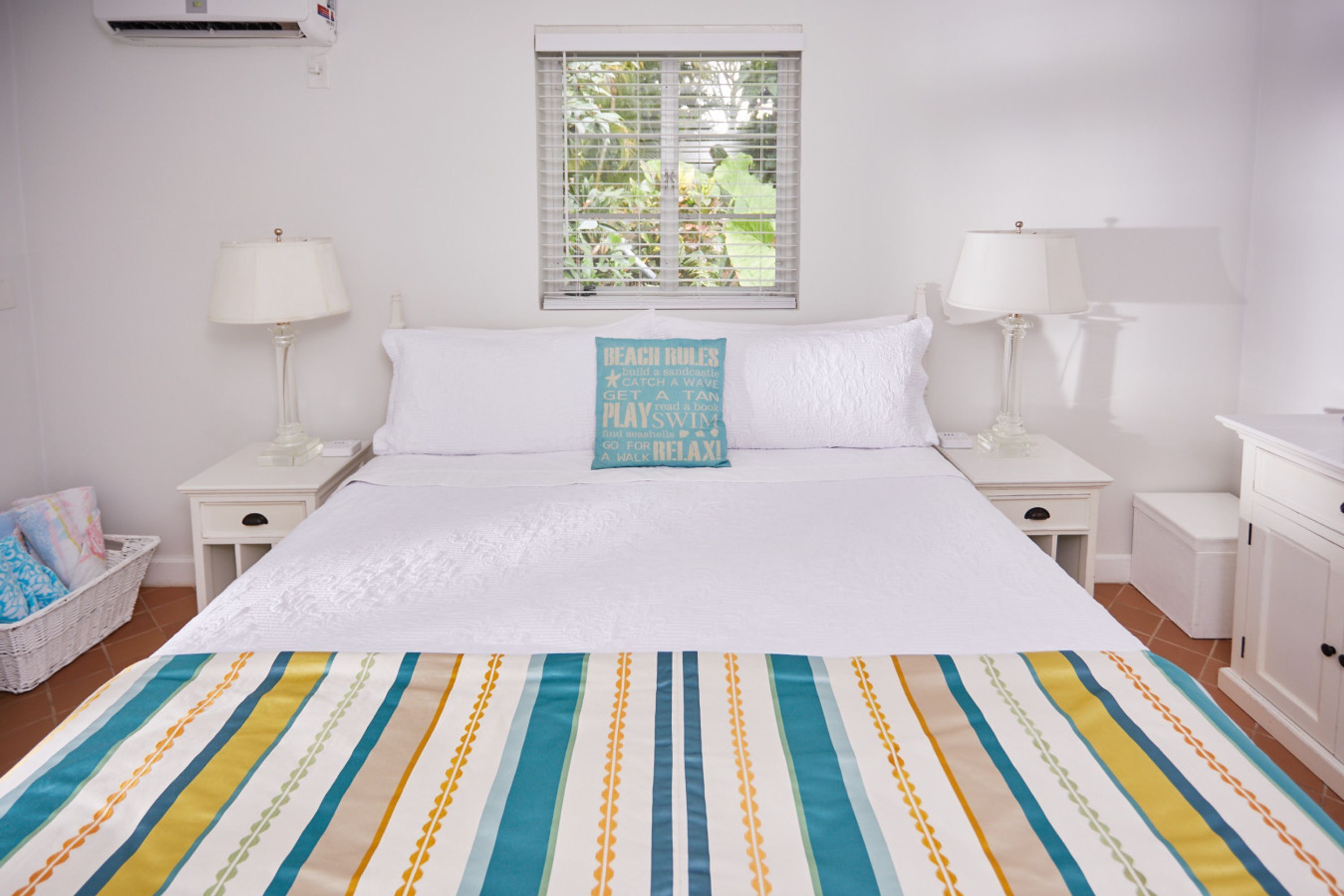 Une chambre à coucher bien aménagée, avec un grand lit, deux tables de chevet, une fenêtre avec des stores et un coussin sur le thème de la plage.