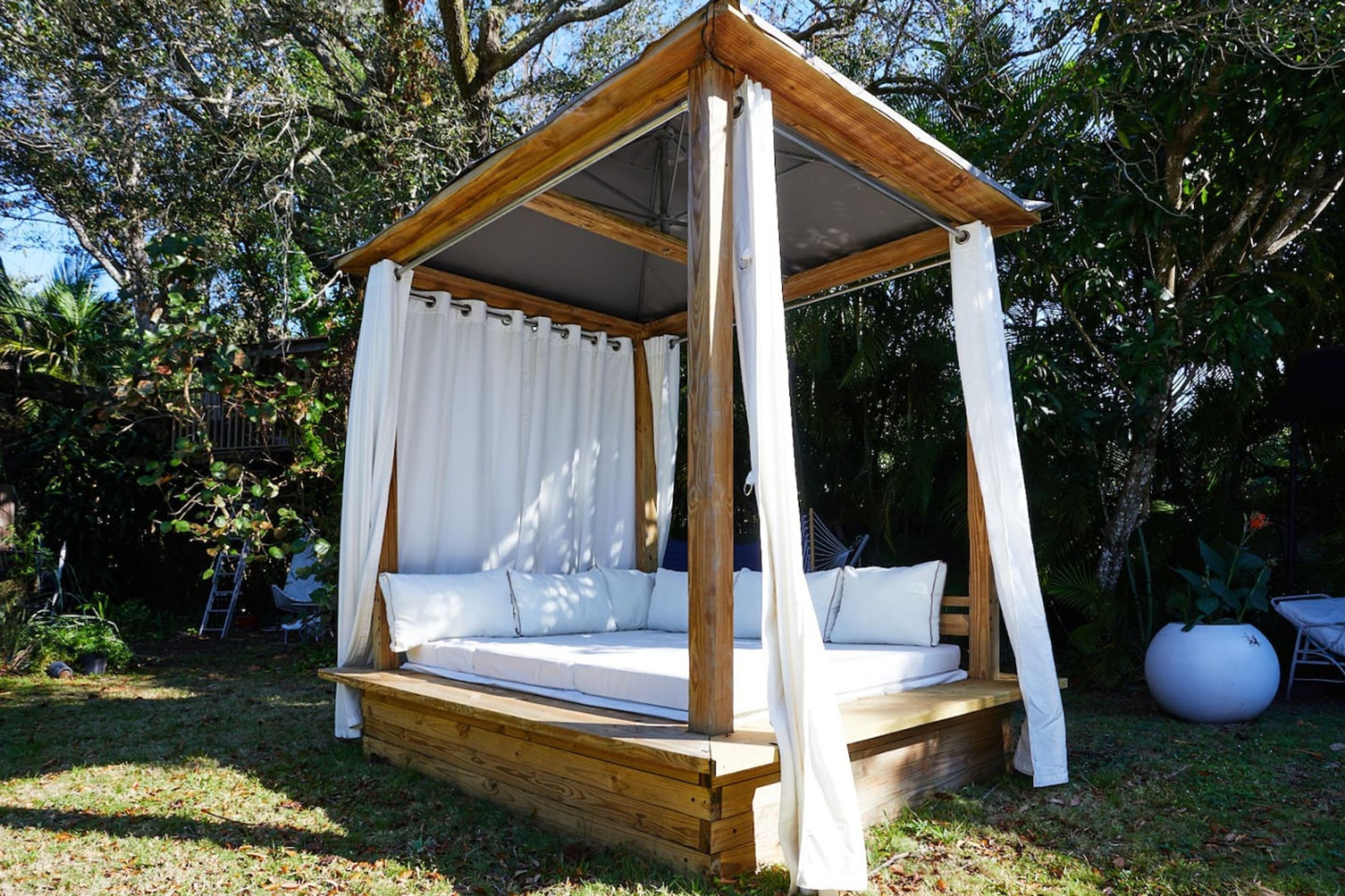 Una cama con dosel de madera y cortinas blancas, ubicada en un jardín con árboles y una maceta blanca al fondo.