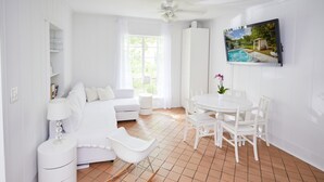 A bright, white room with a sofa, a dining table with chairs, a TV mounted on the wall, and a window with curtains. - Miami Pool Cottage in El Portal (Miami)