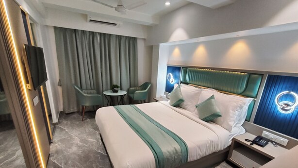 A modern hotel room with a large bed, a desk with a phone, a chair, a small table with a plant, and a ceiling fan. - Hotel Prime Suites Airoli (Navi Mumbai)