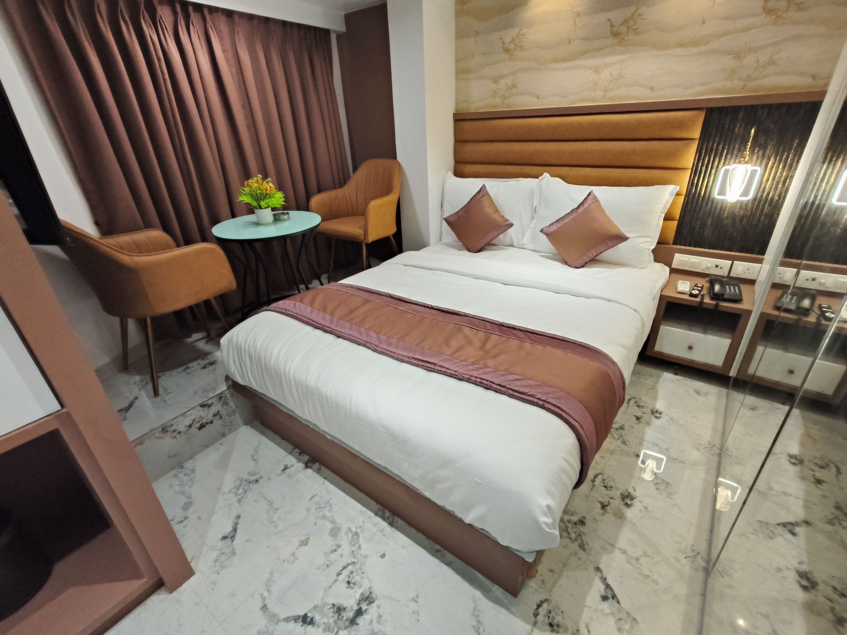 A modern hotel room with a large bed, a small round table with chairs, a nightstand with a phone, and a decorative wall panel.