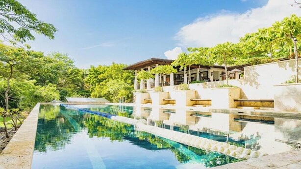 Una piscina con agua cristalina azul, rodeada de exuberante vegetación y una zona de estar cubierta.
