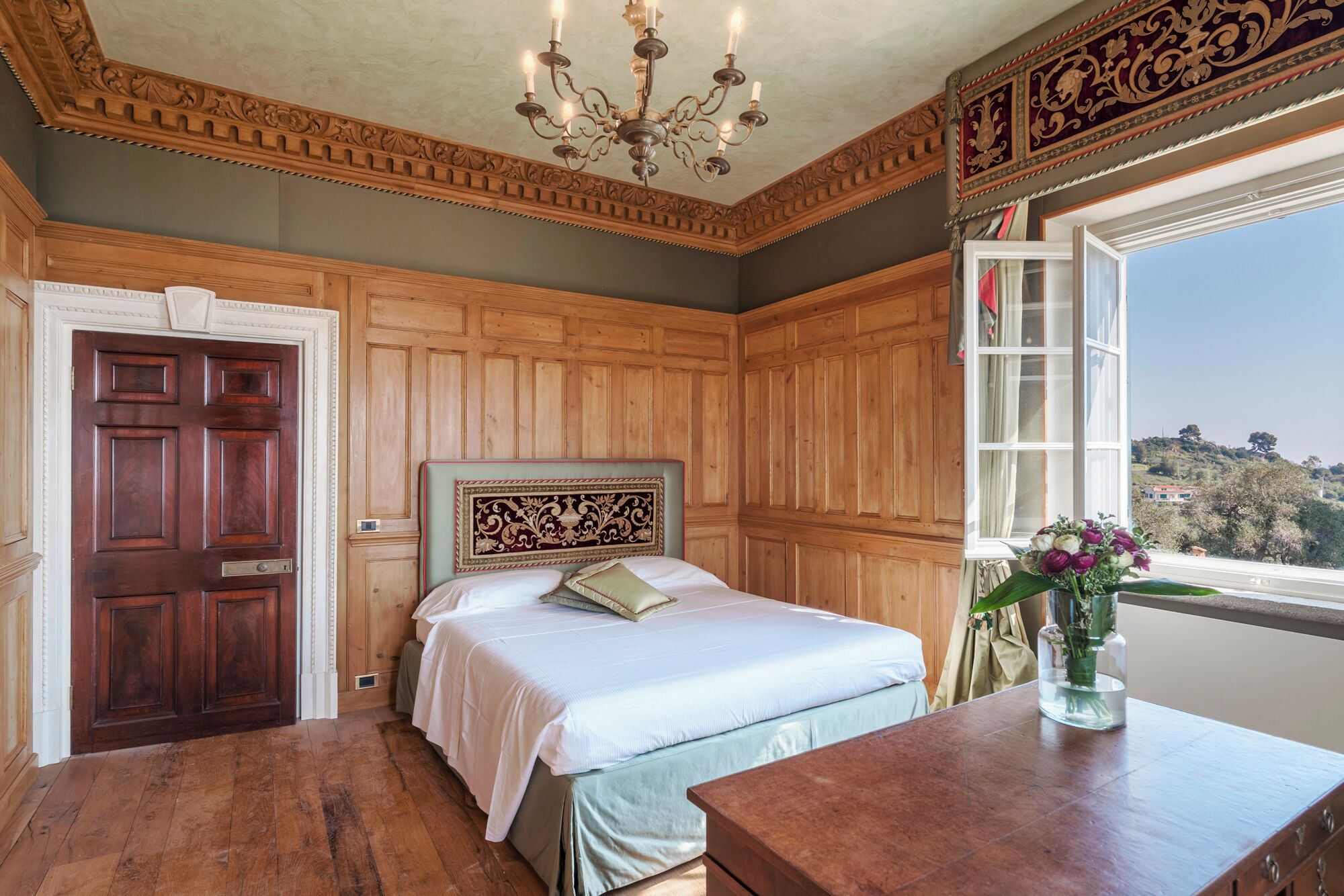 A room with a wooden paneled wall, a bed with a decorative headboard, a wooden table with a vase of flowers, and a window with a view of the landscape.