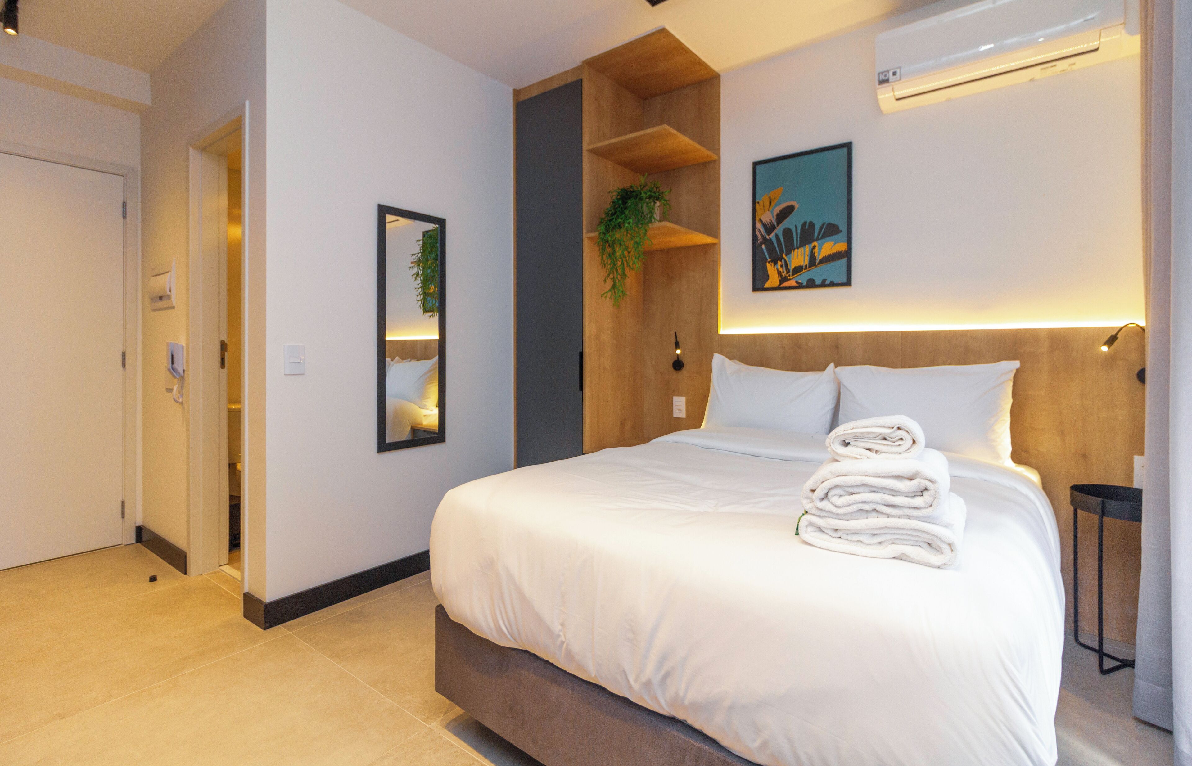 A modern hotel room with a large bed, a wooden headboard, a wall-mounted air conditioner, a framed picture, and a potted plant.