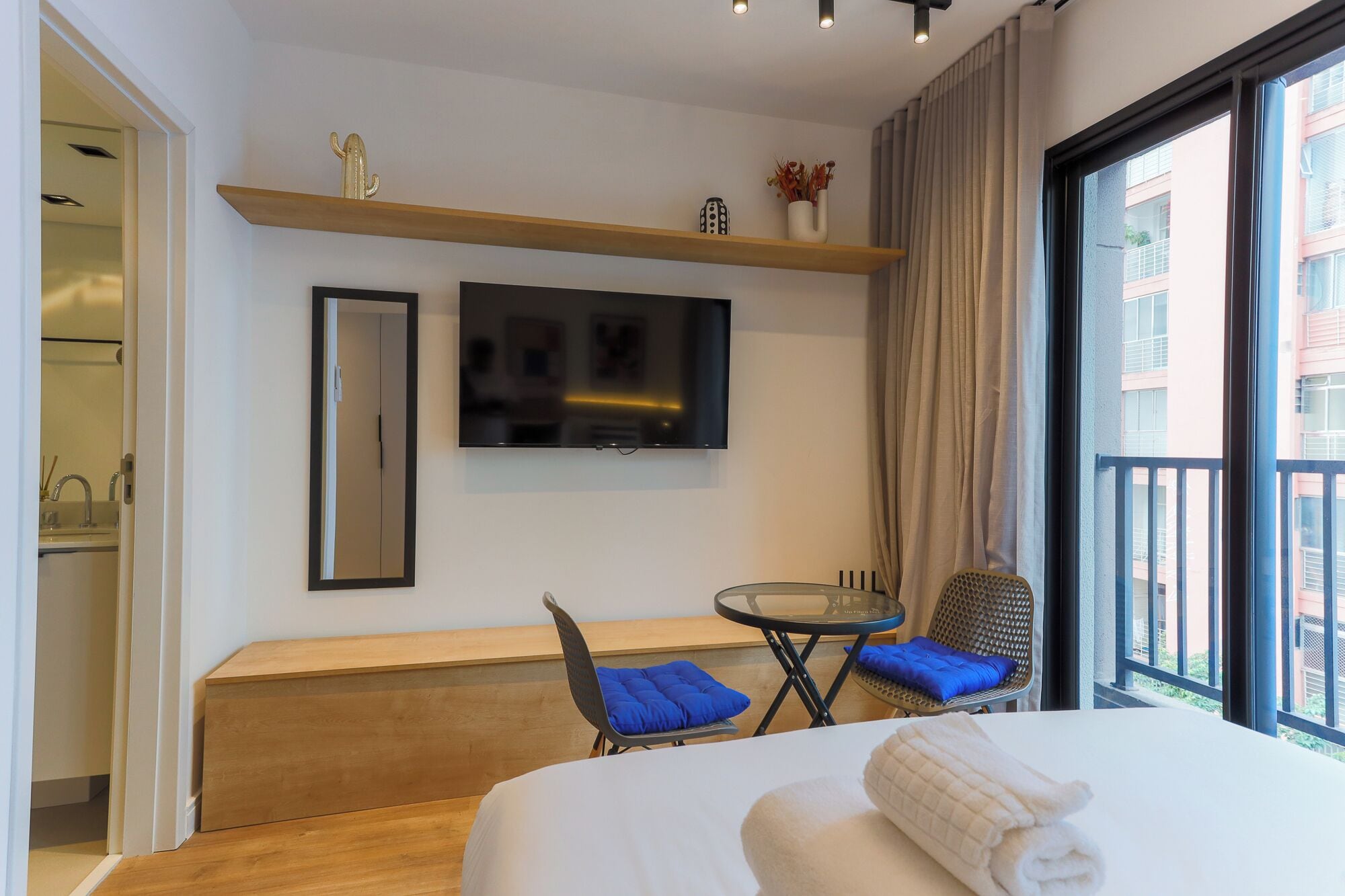 A hotel room with a bed, a TV mounted on the wall, a small table with a chair, a sofa, and a balcony with a view of buildings.