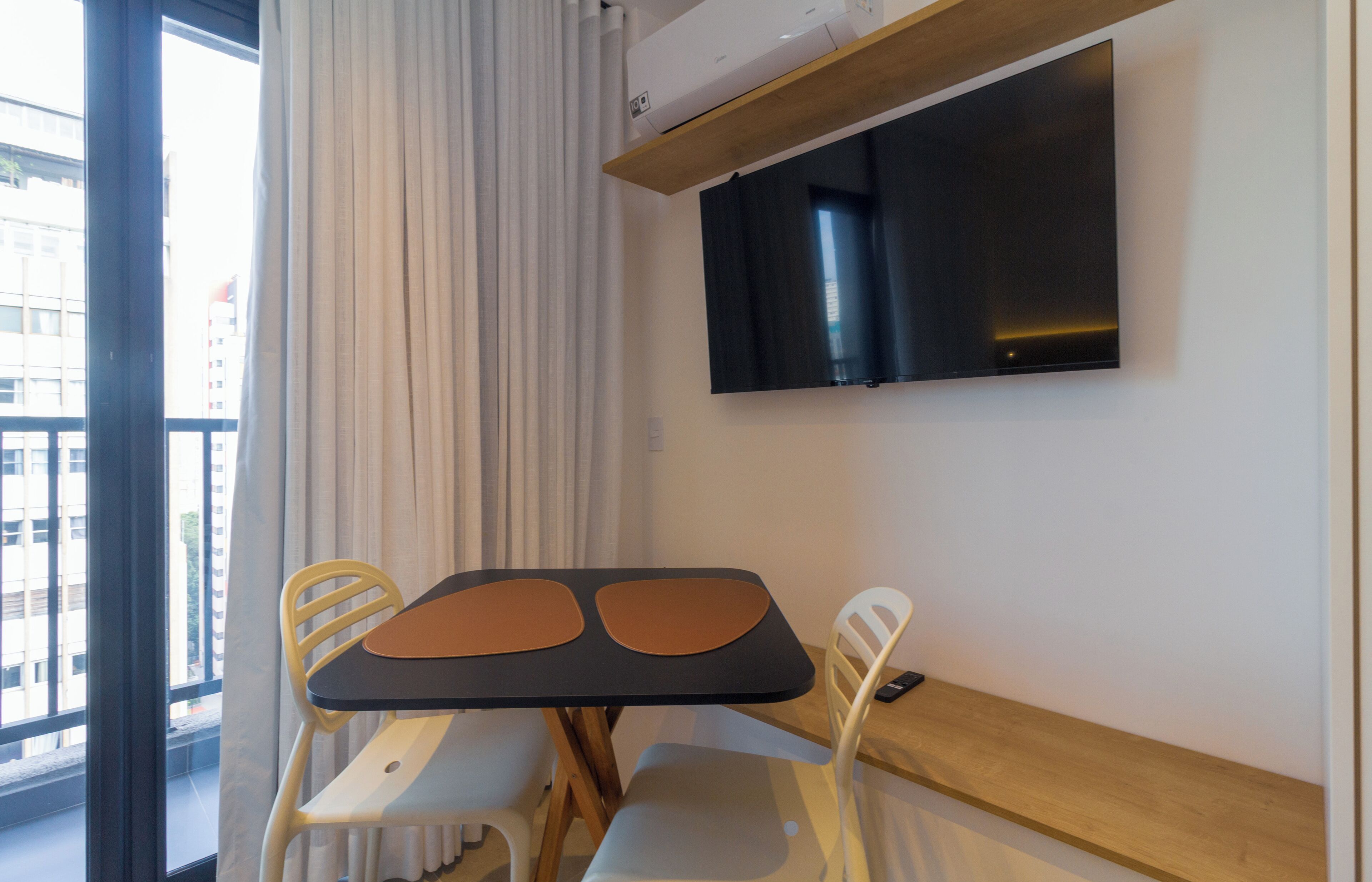 A modern room with a flat-screen TV mounted on the wall, a table with two chairs, and a window with curtains.