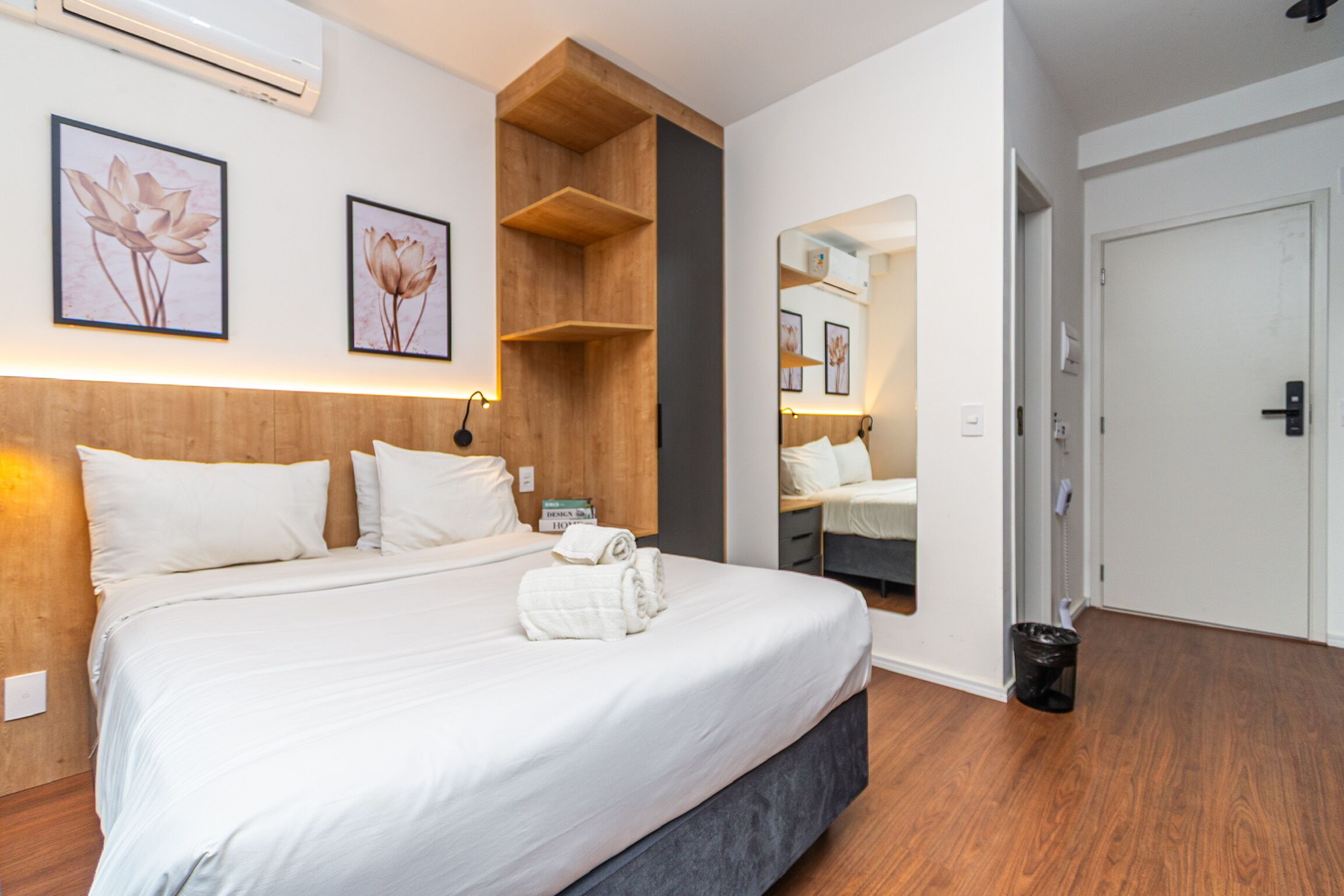 A hotel room with a large bed, wooden headboard, wall-mounted air conditioning, framed floral artwork, a mirror, and a door.