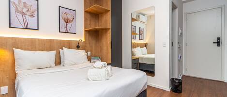 A hotel room with a large bed, wooden headboard, wall-mounted air conditioning, framed floral artwork, a mirror, and a door.