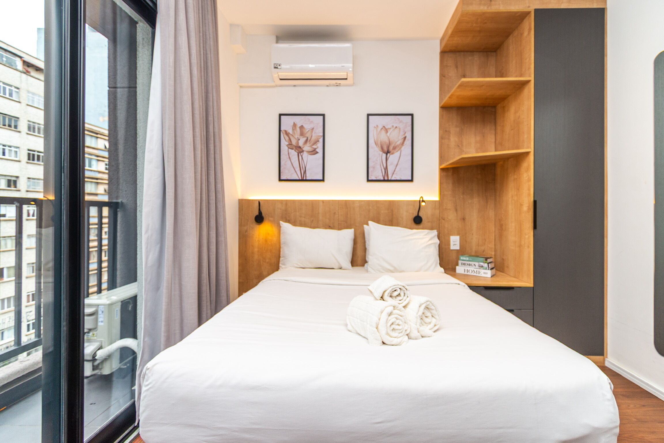 Hotellihuone, jossa on suuri sänky, kaksi kehystettyä kukkaaiheista taulua seinällä, puinen hylly ja ikkuna, jossa on verho.
