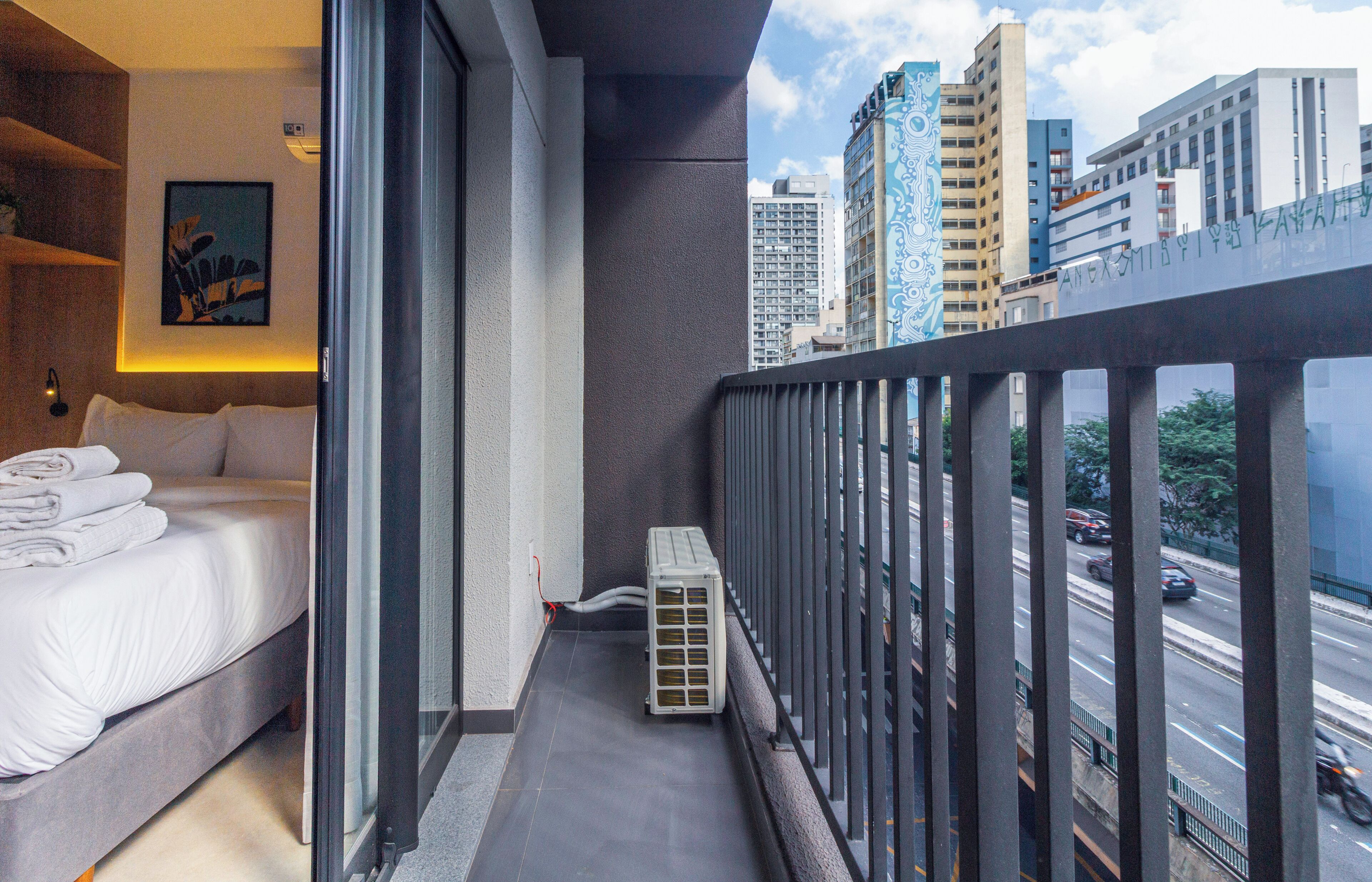 A hotel room with a bed, a radiator, and a view of a cityscape with tall buildings and a busy street.