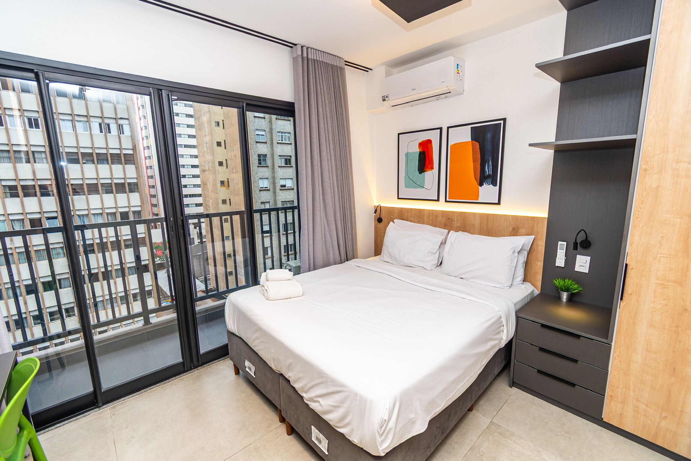Habitación de hotel moderna con una cama grande, un balcón con vistas a edificios, un escritorio pequeño y un aire acondicionado de pared.