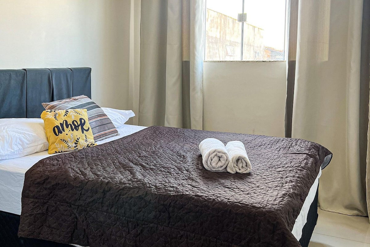 A neatly made bed with a brown quilt, two rolled towels, and a yellow pillow with the word "amor" on a hotel room.