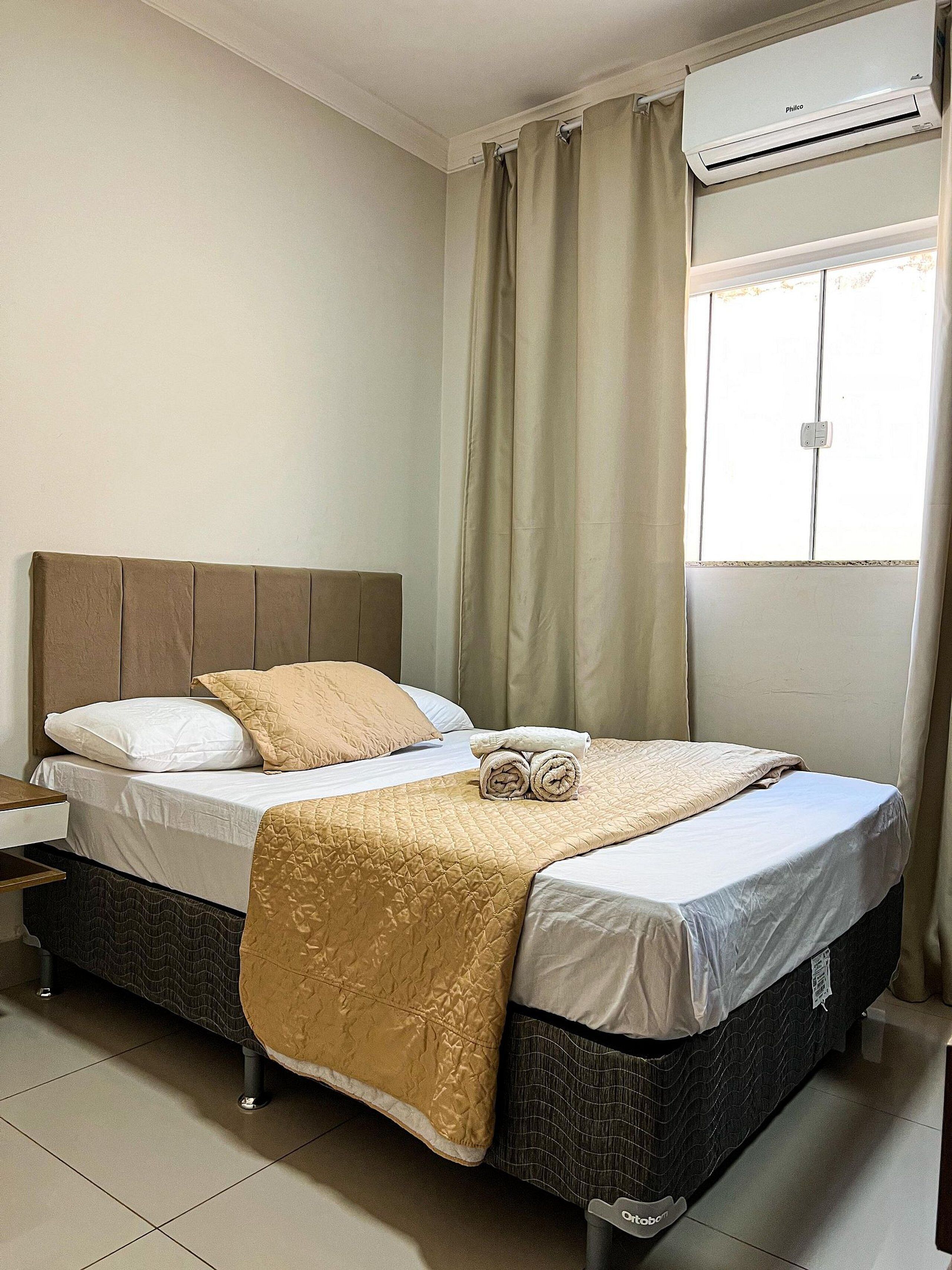 A neatly made bed with a beige quilt, a pillow, and rolled towels. There is a window with curtains, an air conditioning unit, and a nightstand with a lamp.
