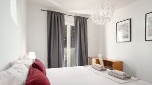 Een moderne slaapkamer met een groot bed, een bureau en een stoel. Er zijn gordijnen bij het raam, een kroonluchter en ingelijste foto's aan de muur.