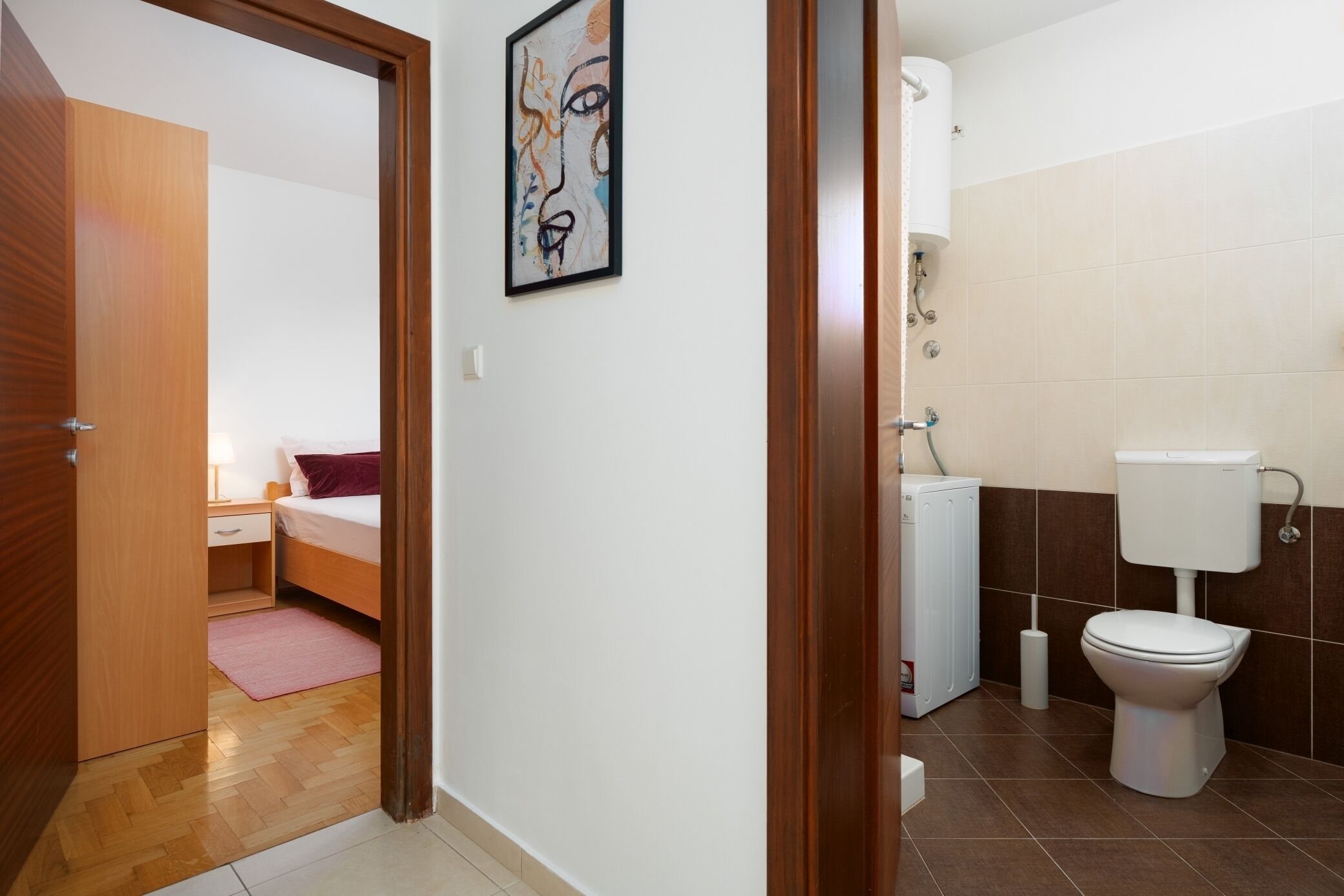 A hotel room with a wooden door, a bed with a pink rug, a white bedside table, and a wall-mounted painting.