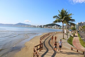 A beach with a wooden boardwalk, palm trees, and a cityscape in the background. - Apê a 50 metros do Mar - Itapema (Itapema)