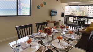Un coin repas avec une table dressée pour quatre personnes, des chaises, un téléviseur fixé au mur et une vue sur la mer par une grande fenêtre.
