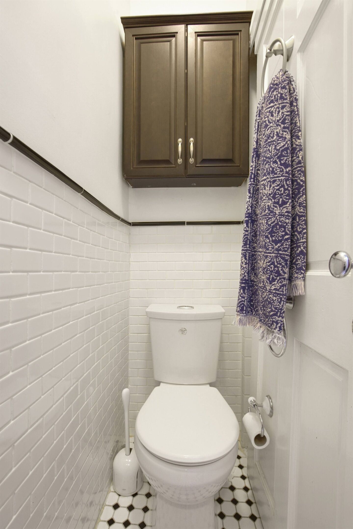 A bathroom with white subway tiles, a white toilet, a brown cabinet, and a patterned towel hanging on the wall.