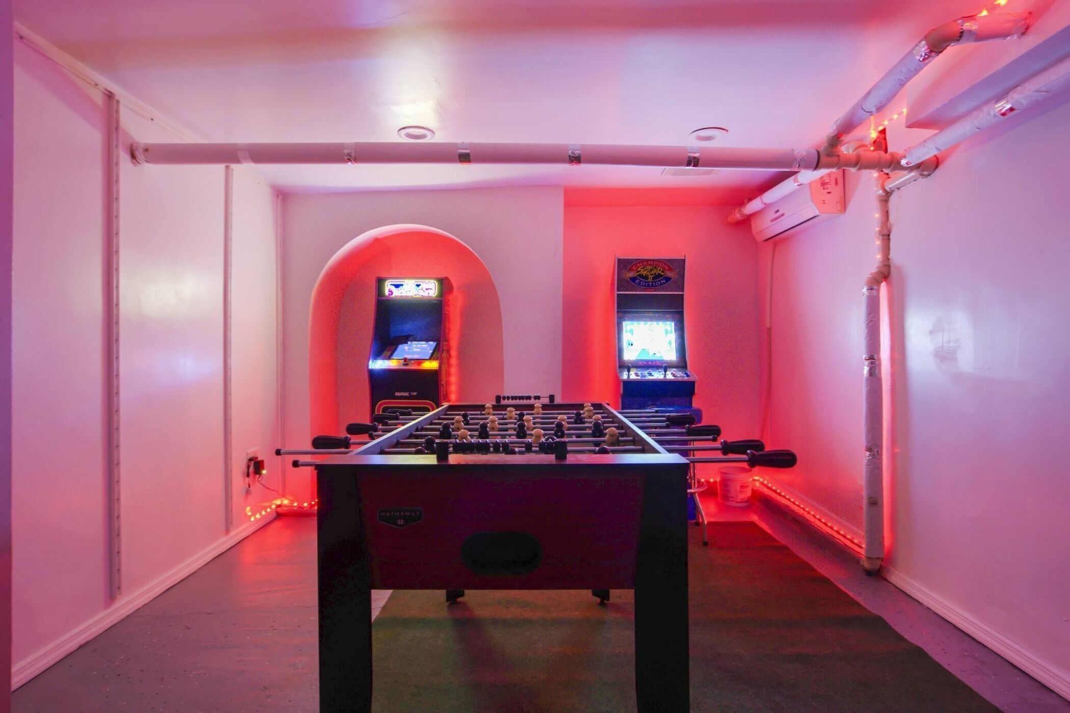 A room with a foosball table, two arcade machines, and a red neon light strip along the wall.