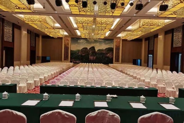 Un salón de conferencias con filas de sillas blancas, un escenario con una mesa verde y un mural grande en la pared de fondo.