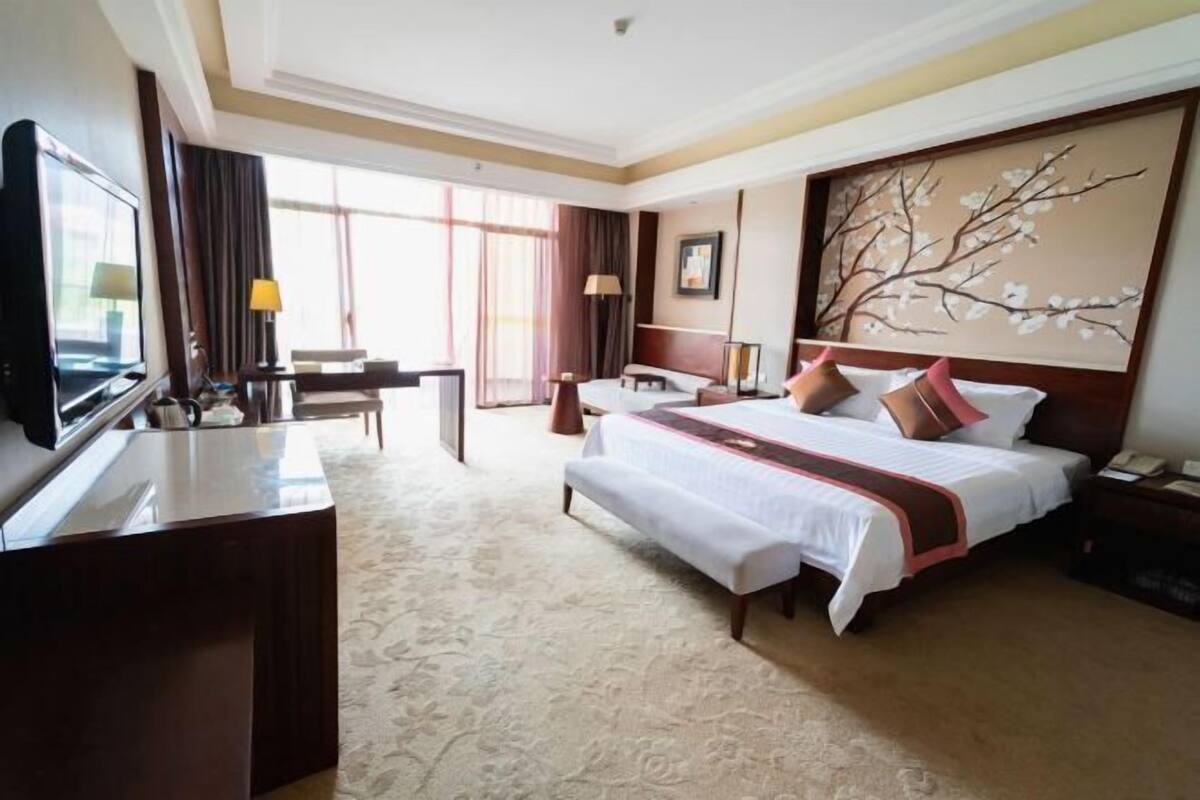 Une chambre d’hôtel avec un grand lit, un bureau avec une télévision, une chaise, une table de chevet et une fenêtre avec des rideaux.