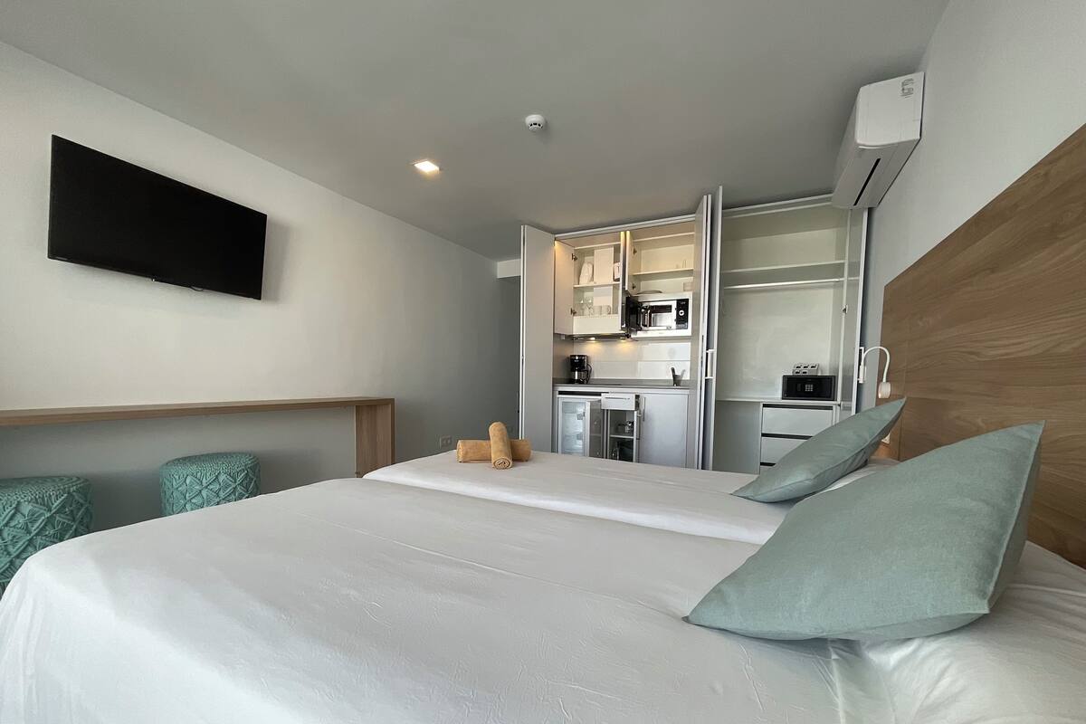 A hotel room with a bed, a TV mounted on the wall, a kitchenette, and a wooden headboard.